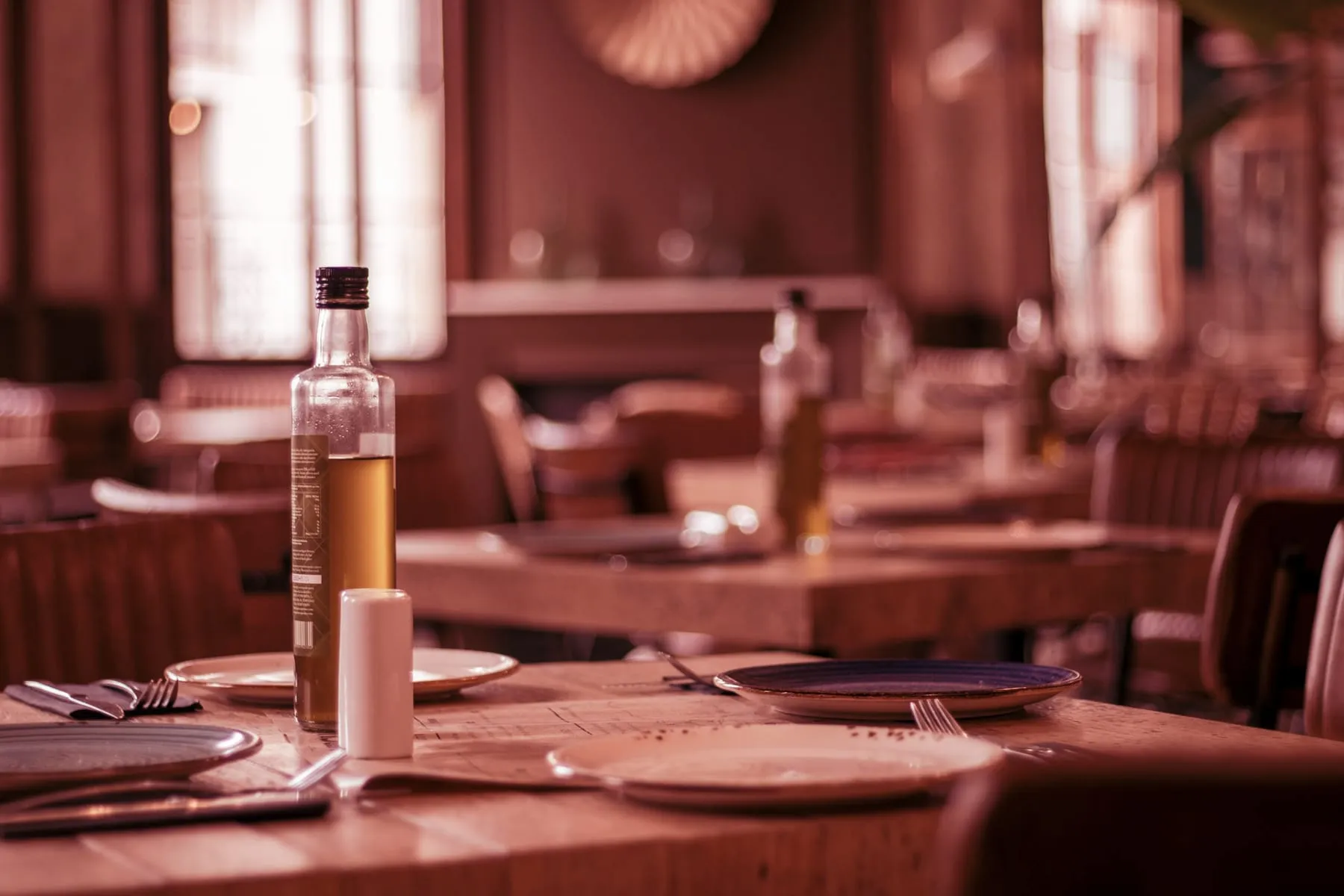 Une table de bistrot dressée au premier plan, bouteille d'huile et assiettes dépareillées, la salle en bois derrière dans la lumière du jour